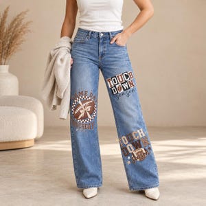 Peut inclure: Un jean en denim bleu porté avec un débardeur blanc et un gilet beige. Le jean est orné de motifs sur le thème du football en marron et blanc, incluant le texte "TOUCHDOWN season" et "GAME DAY TOUCHDAY" avec des illustrations de ballons.