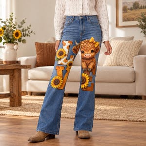Peut inclure: Jean évasé en denim bleu avec un imprimé graphique sur le devant. Le motif comprend le mot LOVE en lettres capitales avec un imprimé vache et tournesol, ainsi qu'un veau highland brun et duveteux portant un nœud jaune sur la tête.