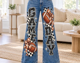May include: Blue denim flare jeans featuring a graphic print with the text GAME DAY in black block letters. The design includes two brown footballs with leopard print bows, black and white checkered patterns, and small star shapes.