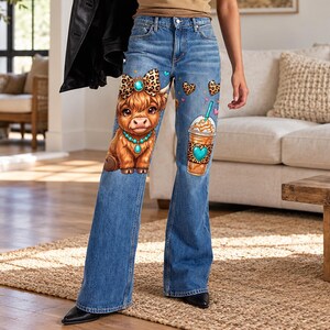 May include: Blue denim flare jeans featuring a graphic print of a brown Highland cow with a leopard print bow and turquoise necklace on the left leg, and an iced coffee drink with a turquoise heart on the right leg, accented by small hearts.