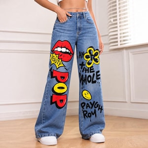Puede incluir: Vaqueros de mezclilla azul de talle alto con perneras anchas y estampados gráficos coloridos. La pierna izquierda muestra un icono de labios rojos con el texto BEAUTY y POP. La pierna derecha presenta una flor amarilla, una cara sonriente y texto.