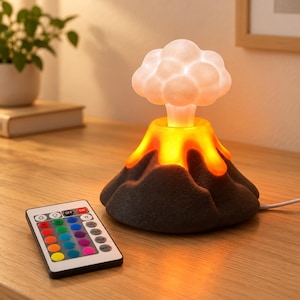 Puede incluir: Una lámpara LED con forma de volcán, base texturizada gris oscuro, efecto de lava naranja brillante y una parte superior blanca similar a una nube. Incluye un mando a distancia rectangular con botones de colores para ajustar la iluminación.