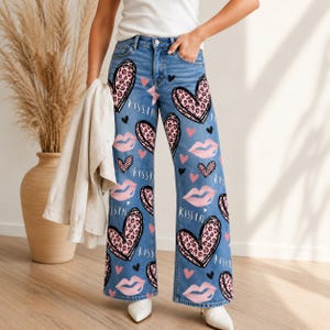 Puede incluir: Vaqueros de mezclilla azul con silueta de pierna ancha y un patrón repetitivo de corazones con estampado de leopardo rosa, labios rosas y la palabra "KISS" en letra blanca. Los pantalones tienen un diseño clásico de cinco bolsillos con cremallera.