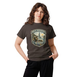 Op de afbeelding: Een donkerbruin t-shirt met korte mouwen en een grafische print van een persoon die stenen zoekt aan een oever met een zeilboot op de achtergrond. Het ontwerp bevat de tekst "ROCK HOUNDING" bovenaan en "DIG DEEPER" onderaan het embleem.