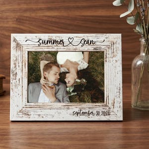 Può includere: Una cornice rettangolare con finitura in legno bianco anticato. Il bordo superiore presenta la scritta nera "summer & Sean" con un simbolo a cuore, mentre il bordo inferiore riporta la scritta nera "september 30.2026". All'interno c'è la foto di una coppia nell'erba.