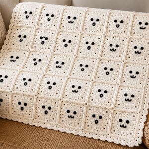 May include: A cream-colored crochet blanket composed of square motifs, each featuring a unique black yarn-stitched facial expression. The blanket has a scalloped border and displays a grid of various ghost-like faces with different eyes and mouths.