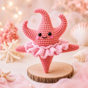 May include: A crocheted pink starfish plush toy featuring a smiling face with black bead eyes and small pink cheeks. The starfish is wearing a light pink ruffled tutu around its center and is positioned upright on a circular wooden base.
