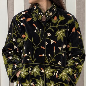 May include: A black velvet jacket featuring a high collar and long sleeves, adorned with intricate embroidery of green leaves and orange and white floral motifs. The jacket includes two front pockets and a button-down closure.