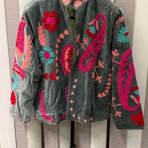 May include: A grey velvet jacket featuring vibrant floral and paisley embroidery in shades of hot pink, turquoise, and red. The jacket has a high collar, long sleeves, and a button-front closure, with the embroidery extending across the front and sleeves.