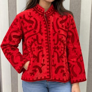 May include: A bright red velvet jacket featuring a mandarin collar and long sleeves. The jacket is adorned with a darker red, raised floral and vine pattern throughout the fabric. It includes a front opening and two side pockets.