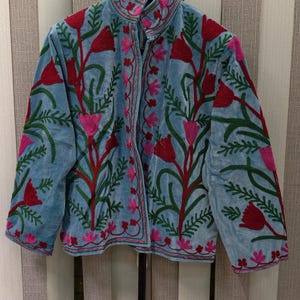 May include: A light blue jacket featuring a high collar and long sleeves, decorated with vibrant red and pink floral embroidery and green leafy stems. The garment is constructed with a front opening and displays intricate needlework patterns across the entire surface, including the sleeves and the front panels.