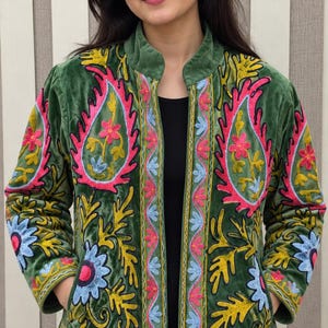 May include: A long-sleeved olive green velvet jacket featuring vibrant, multi-colored floral and paisley embroidery in shades of pink, yellow, and light blue. The jacket has a stand-up collar, an open front, and two side pockets with detailed stitching.