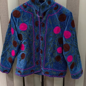 May include: A blue velvet jacket featuring a high collar and long sleeves, decorated with embroidered dark brown and bright pink circular motifs, accented by intricate green leaf patterns and a vibrant pink stitched trim along the edges.
