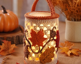 May include: A cylindrical crochet lantern featuring a cream-colored openwork body adorned with appliqued autumn leaves in shades of orange, brown, and red. The lantern has a solid orange crochet handle and a matching orange rim at the top and base.
