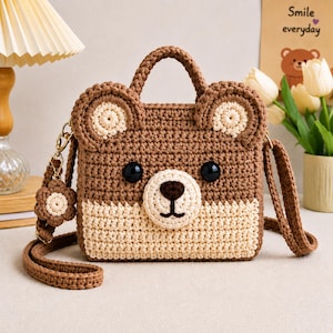 Könnte beinhalten: Eine braun-cremefarbene Häkeltasche in Form eines Bärengesichts mit zwei abgerundeten Ohren, schwarzen Kunststoffaugen und einer gestickten Nase. Die Tasche verfügt über einen Tragegriff, einen langen Schulterriemen und einen kleinen blumenförmigen Anhänger.