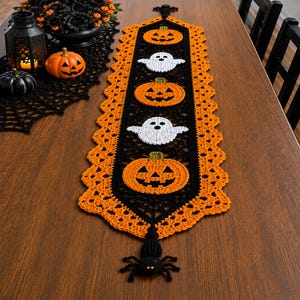 May include: A long, rectangular crochet table runner featuring a black centre panel with three orange jack-o'-lanterns and two white ghosts. The runner has a wide, scalloped orange border and a black crocheted spider at each pointed end.