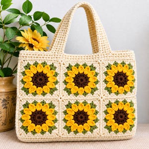 May include: A cream-coloured crochet tote bag featuring six square panels, each displaying a sunflower motif with a dark brown centre, bright yellow petals, and green leaves. The bag has two sturdy handles and a structured rectangular shape.