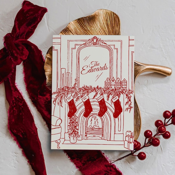 May include: A cream Christmas card featuring a red illustration of a fireplace, stockings, and the text "The Edwards." The card rests on a gold tray, with a red velvet ribbon and red berries.