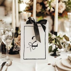 Pode incluir: Um cartão de número de mesa preto e branco com um laço de fita preto e o número "2" impresso nele. O cartão está sobre uma toalha de mesa branca.