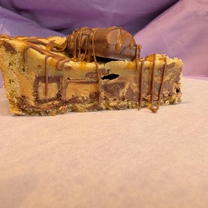 May include: A thick slice of golden-brown cookie pie featuring a dense, textured interior with visible chocolate chunks and caramel swirls. The top is garnished with a whole chocolate bar piece and drizzled with thin lines of melted chocolate.