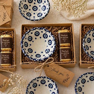 Puede incluir: Varios cuencos pequeños de cerámica con un patrón de ojo turco azul y blanco, empaquetados en cajas de cartón marrón con relleno de papel, dos chocolates envueltos en oro y una etiqueta de papel marrón con el texto Thank you.