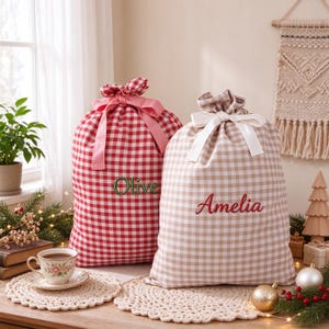 May include: Two fabric drawstring gift bags featuring a gingham pattern. The left bag is red and white with a pink ribbon and the name Olive embroidered in green. The right bag is beige and white with a white ribbon and the name Amelia in red.