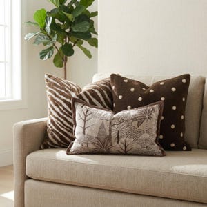 May include: Three decorative throw cushions arranged on a sofa. One cushion features a brown and white zebra stripe pattern, another is a dark brown square with white polka dots, and the third is a rectangular cushion with a brown botanical print.