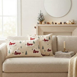 May include: Two rectangular throw pillows with an off-white fabric base featuring embroidered red houses with white snow roofs and green pine trees. The pillows are arranged on a light beige sofa, showcasing a repeating winter village pattern.