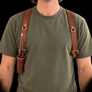 Könnte beinhalten: Ein Schultergurt aus braunem Leder mit verstellbaren Riemen, messingfarbenen Metallbeschlägen und einer Holstertasche auf der rechten Seite, in der sich eine Metallpinzette befindet. Der Gurt wird über einem olivgrünen T-Shirt getragen.