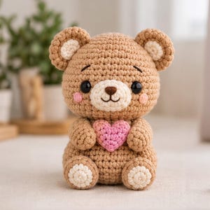 Puede incluir: Un oso de peluche tejido a ganchillo en color marrón claro con hocico, interior de las orejas y almohadillas de color crema. El oso tiene ojos de plástico negro, nariz bordada en marrón y mejillas rosadas. Sostiene un corazón rosa texturizado.