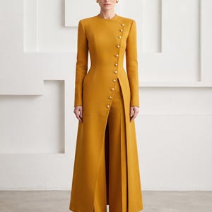 Puede incluir: Una chaqueta larga de color amarillo mostaza con cuello alto y una hilera diagonal de botones dorados que va desde el cuello hasta la cintura, combinada con pantalones anchos a juego. La chaqueta tiene mangas largas y silueta estructurada.
