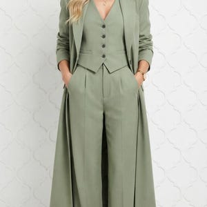 May include: A sage green three-piece suit consisting of a long, open-front blazer, a button-up vest, and wide-leg trousers with a pleated front. The ensemble features a consistent muted green tone throughout all three tailored garments.