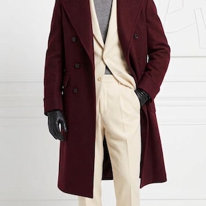 Op de afbeelding: Een bordeauxrode wollen overjas, een crèmekleurig colbert en pantalon, een grijze coltrui, een bijpassende bordeauxrode fedora hoed, zwarte leren handschoenen en bordeauxrode loafers met kwastjes. Het ensemble is voorzien van gestructureerd maatwerk.