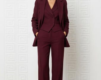 Könnte beinhalten: Ein tiefburgunderfarbener Dreiteiler, bestehend aus einem taillierten Blazer, einer zweireihigen Weste mit Knöpfen und einer passenden Hose mit weitem Bein. Das Ensemble zeichnet sich durch einen strukturierten Kragen und lange Ärmel aus.