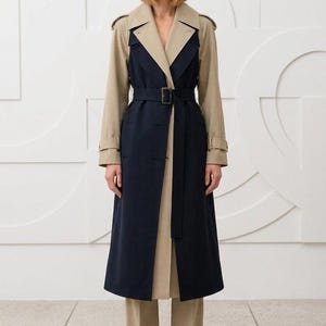 May include: A long trench coat featuring a color-blocked design with a dark navy blue central panel and beige sleeves, collar, and back. The coat includes a matching fabric belt with a rectangular buckle, shoulder epaulets, and beige trousers.