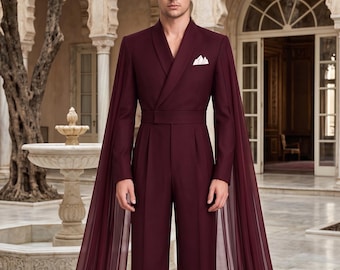 May include: A deep burgundy jumpsuit featuring a shawl collar, a fitted waist, and long, flowing sheer cape sleeves that extend to the floor. The garment includes a white pocket square tucked into the chest pocket and wide-leg trousers.