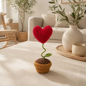 May include: A crocheted heart-shaped flower in a small brown pot. The heart is bright red with a textured yarn surface. A thin, winding green stem connects the heart to the soil, featuring a single small green leaf attached to the stem.