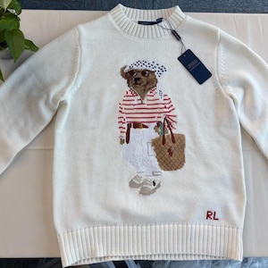 May include: A cream-colored long-sleeve knit sweater featuring a central graphic of a bear wearing a white and black patterned headscarf, sunglasses, a red and white striped shirt, white pants, and carrying a brown tote bag with sneakers.