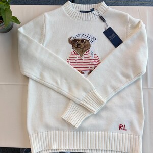 May include: A cream-colored long-sleeve knit sweater featuring a central graphic of a bear wearing a white and black patterned headscarf and a red and white striped shirt. The sweater has a ribbed crew neckline, cuffs, and hem, with RL in red.
