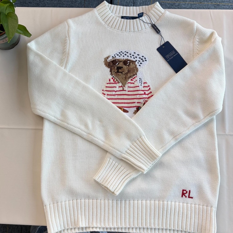 May include: A cream-colored long-sleeve knit sweater featuring a central graphic of a bear wearing a white and black patterned headscarf and a red and white striped shirt. The sweater has a ribbed crew neckline, cuffs, and hem, with RL in red.