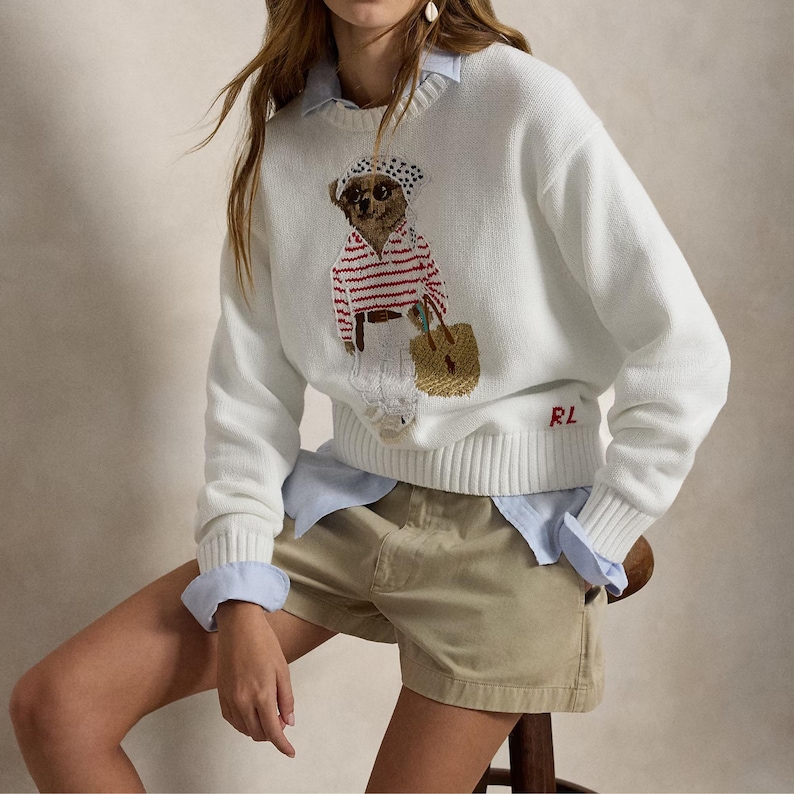 May include: A white long-sleeve knit sweater featuring a graphic of a bear wearing a polka dot headscarf, a red and white striped shirt, and white pants while carrying a tan woven bag. The letters RL are embroidered in red on the lower left side.
