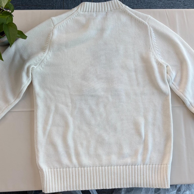 May include: A solid white long-sleeve sweater featuring a ribbed crew neckline, ribbed cuffs, and a ribbed hem. The garment is constructed with a knit texture and includes cable knit detailing along the raglan sleeve seams on the front.