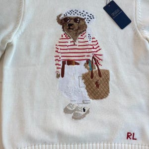 May include: A cream-colored knit sweater featuring a central graphic of a brown bear wearing a white and red striped shirt, white pants, a patterned headscarf, and sunglasses, carrying a tan tote bag. The letters RL are embroidered in red.