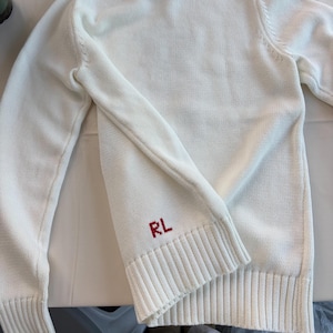 May include: A white long-sleeve knit sweater featuring a ribbed crew neckline, ribbed cuffs, and a ribbed hem. The lower front section of the sweater displays the letters RL embroidered in red thread, positioned just above the ribbed hemline.