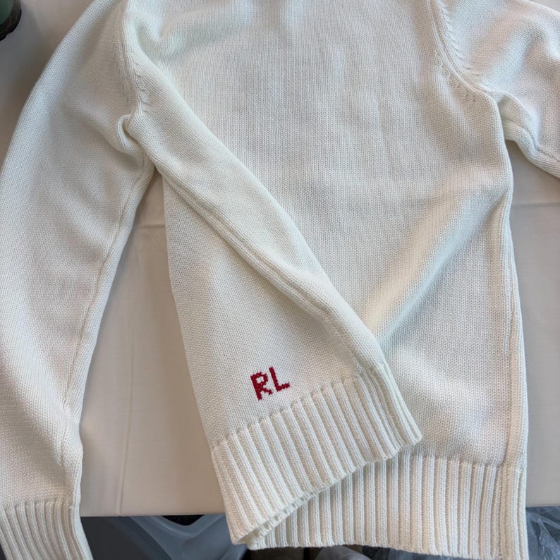 May include: A white long-sleeve knit sweater featuring a ribbed crew neckline, ribbed cuffs, and a ribbed hem. The lower front section of the sweater displays the letters RL embroidered in red thread, positioned just above the ribbed hemline.