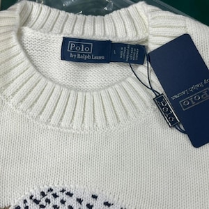 May include: A white knit sweater with a ribbed crew neckline. A navy blue brand label reads Polo by Ralph Lauren with a size L indicator. A matching navy blue rectangular hang tag is attached with a black string, featuring the Polo brand name.