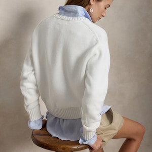 May include: A white long-sleeved knit sweater with a ribbed crew neckline, cuffs, and hem, layered over a light blue collared button-down shirt. The outfit is paired with beige shorts, showcasing a classic layered look with visible shirt cuffs.