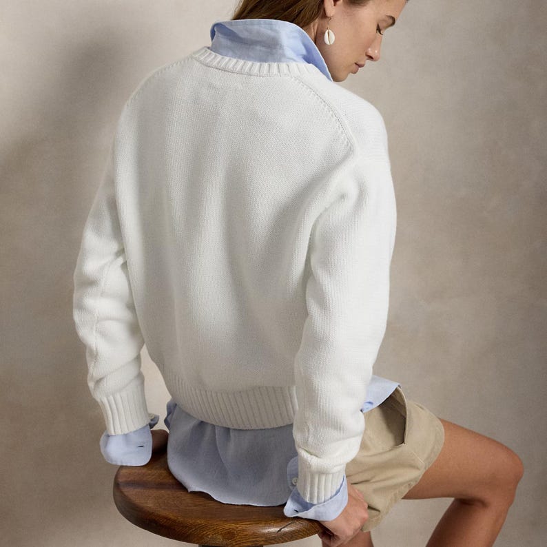 May include: A white long-sleeved knit sweater with a ribbed crew neckline, cuffs, and hem, layered over a light blue collared button-down shirt. The outfit is paired with beige shorts, showcasing a classic layered look with visible shirt cuffs.