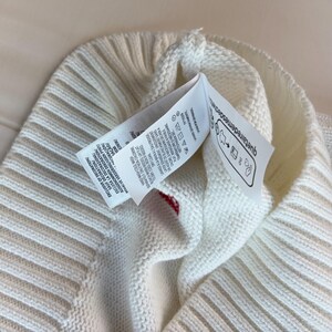 May include: A white knitted beanie with a ribbed cuff and a prominent care label attached to the interior. The label features printed text in multiple languages, laundry care symbols, and the website text quetairedemesdechets.fr.