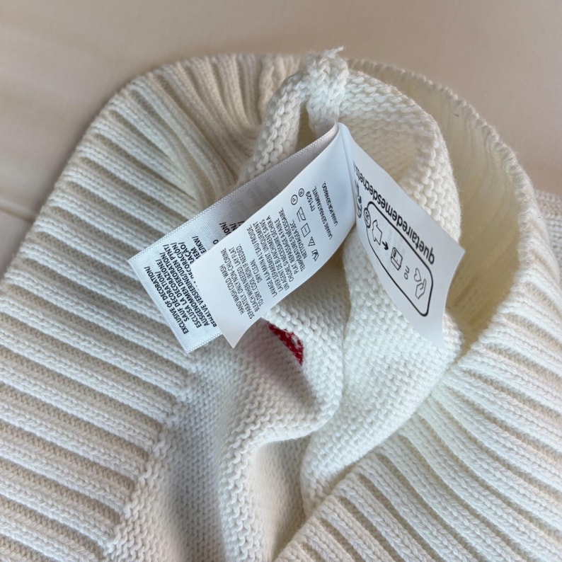 May include: A white knitted beanie with a ribbed cuff and a prominent care label attached to the interior. The label features printed text in multiple languages, laundry care symbols, and the website text quetairedemesdechets.fr.